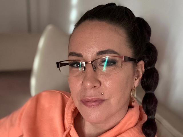 Greekmadame1-hot has dark brown braided hair, glasses, a nose piercing, and is wearing a peach hoodie. She's smiling with light makeup and gold hoop earrings.