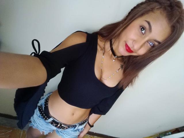 NathaDulcet, com batom vermelho, cropped preto, shorts de jeans e um cinto, tirando uma selfie. Ela tem um sorriso adorável, cabelo castanho e um colar preto..