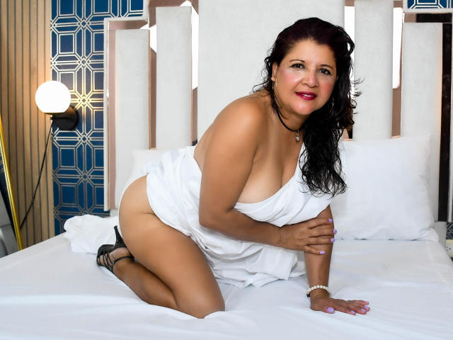 LettieFlores, a curvy woman with dark hair, kneels on a bed, her white dress barely covering her tits and ass, smiling, wearing black heels and a black choker.