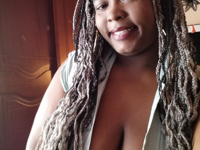 KarlaFlorez has dark brown skin, long blonde and brown braids, and a smile showing her teeth. She's wearing a greenish shirt, revealing her large, round boobs.
