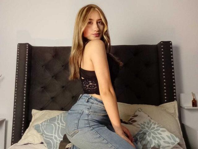 SophiaPaz has blonde hair, a black lace top, and blue jeans, sitting on a bed with a dark, studded headboard. She's looking over her shoulder, showing her ass.