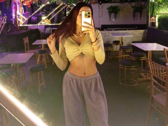 AmberGreyy, con cabello rojo largo, vistiendo una camiseta amarilla corta que deja ver sus pechos y pantalones grises, tomando una selfie frente a un espejo en un bar con poca luz..