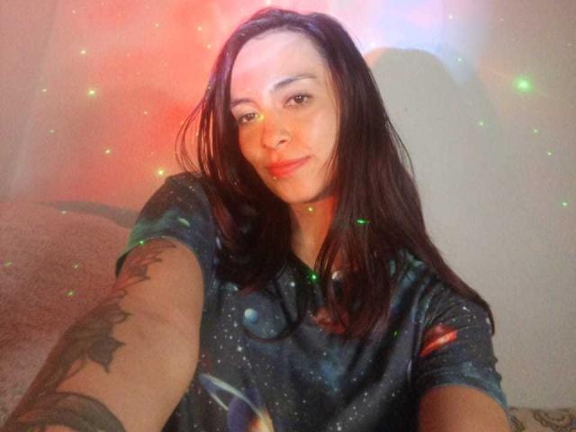 AmberGreyy with long brown hair, wearing a galaxy-patterned shirt, smiling, tattoo on right arm, colorful light reflections, taking a selfie.