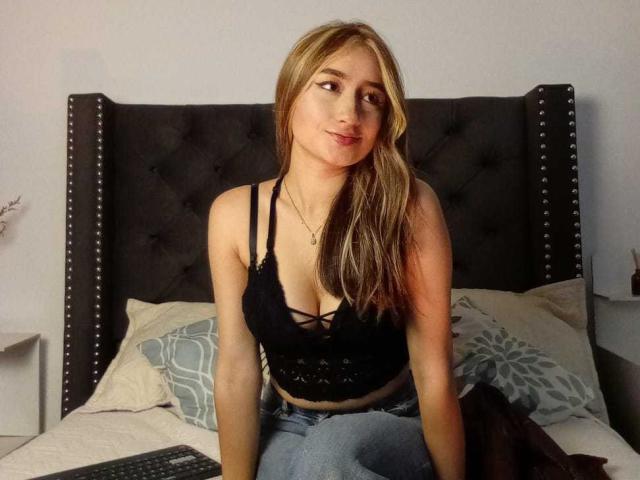 SophiaPaz, con cabello rubio, una blusa de encaje negro que deja ver sus senos, sentada en una cama con un cabecero oscuro y adornado con tachuelas, y almohadas grises, vestida con jeans azules..