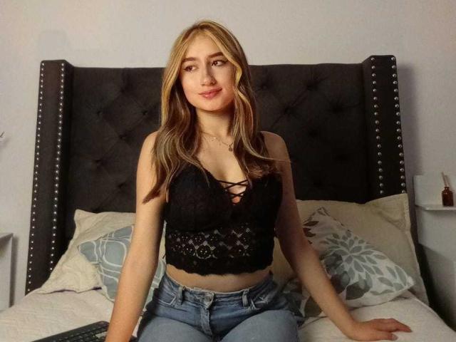 SophiaPaz, com cabelo loiro, blusa de renda preta que revela seios pequenos, e jeans azuis, sentada em uma cama com uma cabeceira escura e com tachas, sorrindo gentilmente..
