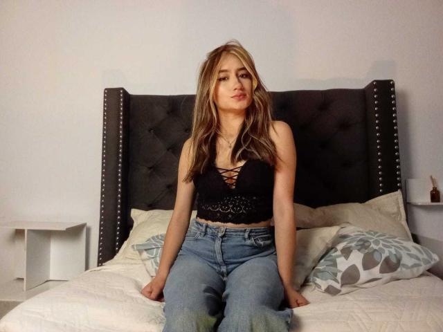 SophiaPaz, com cabelo loiro, usando uma blusa cropped preta com cadarços, que mostra os seios, sentada em uma cama vestindo jeans azuis, olhando para a câmera com um leve sorriso..