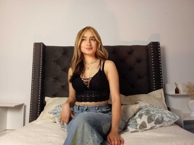 Sophia Paz mit blonden Haaren, einem schwarzen Schnür-BH, der ihre kleinen Brüste zeigt, und Jeans, sitzt auf einem Bett mit einem schwarzen, gepolsterten Kopfteil..