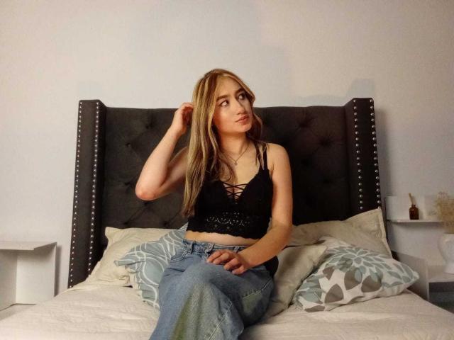 SophiaPaz, avec ses longs cheveux blonds, son crop top noir à lacets et son jean bleu, est assise sur un lit avec une tête de lit grise ornée de clous, et regarde sur le côté, une main dans ses cheveux..