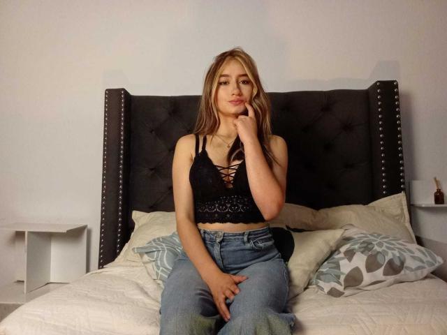 SophiaPaz, con cabello rubio largo, un top corto de encaje negro que deja ver sus pechos y jeans azules, sentada en una cama con una mano en la barbilla, luciendo adorable..