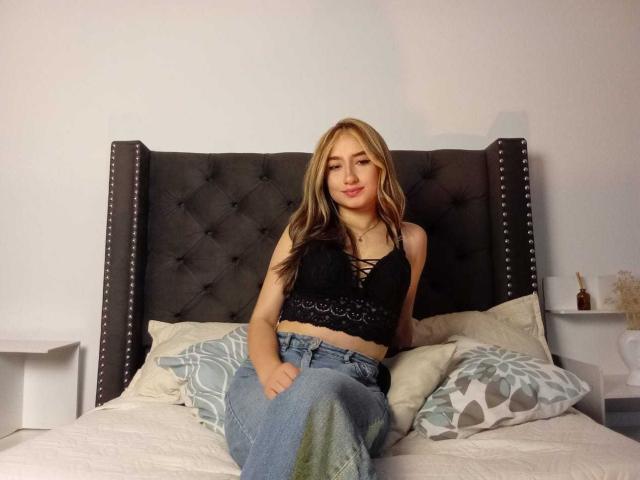 Sophia Paz mit blonden Haaren, die ein schwarzes Spitzen-BH-Top und blaue Jeans trägt, sitzt auf einem Bett mit einem dunkelgrauen, gepolsterten Kopfteil und blickt in die Kamera..
