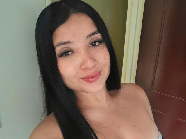 LunaBellaHotty with long black hair, brown eyes, and a slight smile, showing her shiny pink lips and bare shoulders. She's a cute chick with a pretty face.