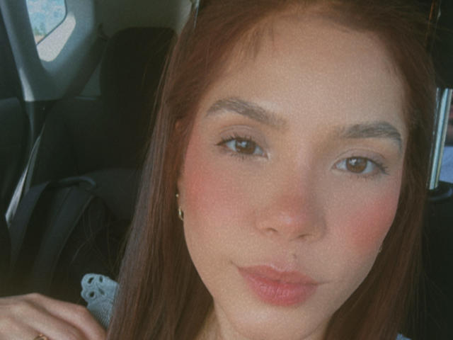 Zyra69 has light brown eyes, flushed pink cheeks, and pouty pink lips, with long brown hair and small gold earrings, sitting in a car. Her right hand is visible.