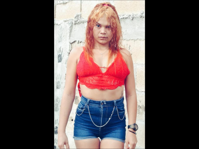 DomiEvans a les cheveux orange et qui ont l'air mouillés, un haut en dentelle rouge et des shorts en jean bleu avec une ceinture à chaîne. Elle a l'air en colère, elle porte une montre et on peut voir un tatouage..