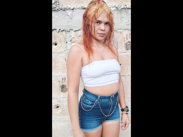 DomiEvans with wet red-orange hair, white tube top, showing her tits, blue denim shorts with chain detail, standing against a brick wall. She's wearing a black wristwatch.