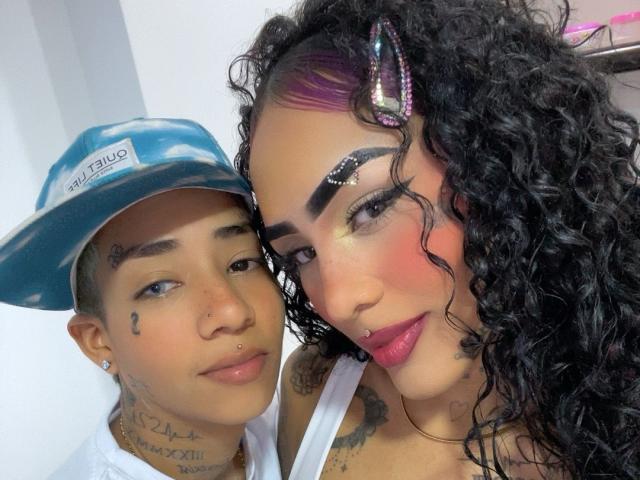 FretcyAmber with curly black hair and purple highlights, wearing pink lipstick, nose ring, and glittery eyeshadow, next to FretcyAmber with a blue cap, tattoos, and a white tank top.