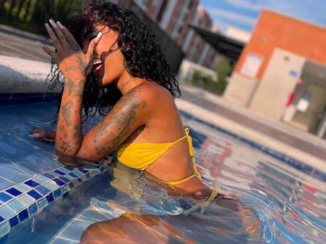 FretcyAmber with tattoos, yellow bikini, black curly hair, hands over face, in pool, water up to her ass, sunlit, blue-tiled edge, urban background, clear sky.