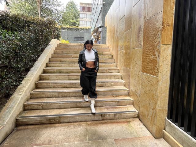 Amberv with dark skin, black hair in a ponytail, white crop top, black jacket, black pants, white socks, and black shoes, standing on stone stairs, hands in pockets.