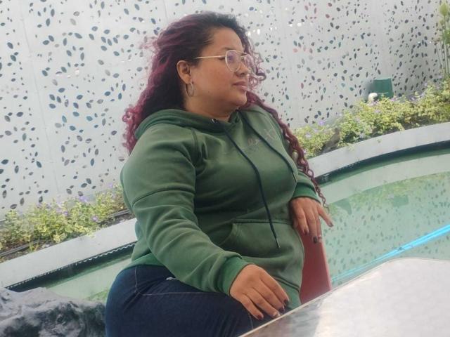 Lalitzfet, met een rond lichaam, grote borsten en een vollere achterkant, draagt een bril, een groene hoodie en donkere jeans, en staat bij een klein zwembad met groen water..
