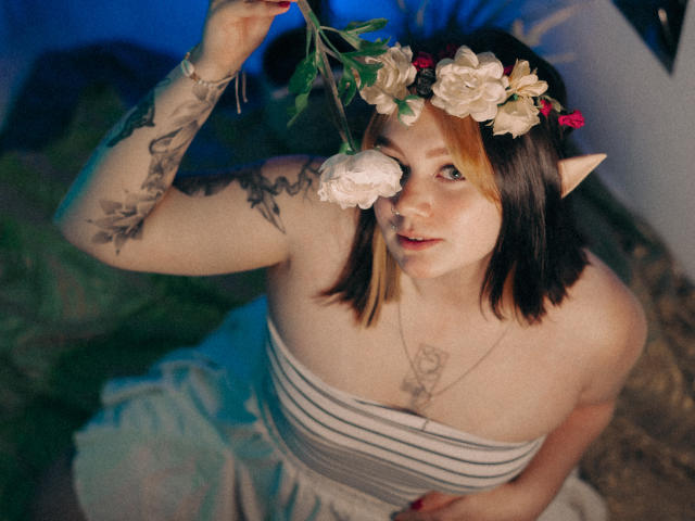 LunaLise with pale skin, brown and blonde hair, elf ears, floral headpiece, tattoos, striped dress, holding flower to nose, looking up, soft lighting, cute, slightly chubby.