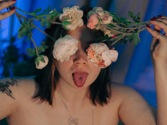 LunaLise with a flower crown, her tongue out, showing a tongue piercing. She has dark hair, tattoos, and is topless, with white and pink flowers in her hands.