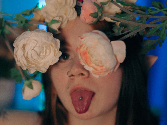 LunaLise with a flower crown, roses covering her eyes, nose piercing, and tongue out with a tongue piercing, blue-lit background.