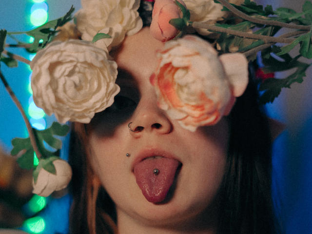 LunaLise with a flower crown, nose ring, and tongue piercing, tongue out, small metal stud in upper lip, dark hair, white and pink roses covering eyes.