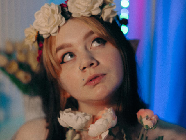 LunaLise has blonde and brown hair, a white flower headband, nose ring, and lip piercing, looking up with blue eyes, surrounded by pink and white flowers.