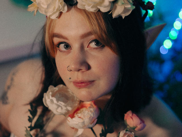 LunaLise with pointed ears, nose ring, and lip piercing, wearing a flower headpiece and necklace, has blonde and black hair, and a cute, slightly freckled face.