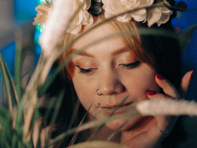 LunaLise with light red hair, floral headpiece, nose ring, and lip piercing, eyes closed, red nails, touching face, soft-focus grass in foreground, blue light from side.