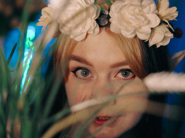 LunaLise with wide blue eyes, wearing a white flower crown, looking through green grass with a slightly open mouth, showing red lips. Blonde and black hair, blue background.