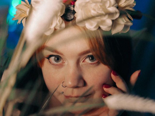 LunaLise with a nose ring, wearing a floral headpiece, has red nails and a subtle smile, with blurred green and white grass in the foreground. She has fair skin.