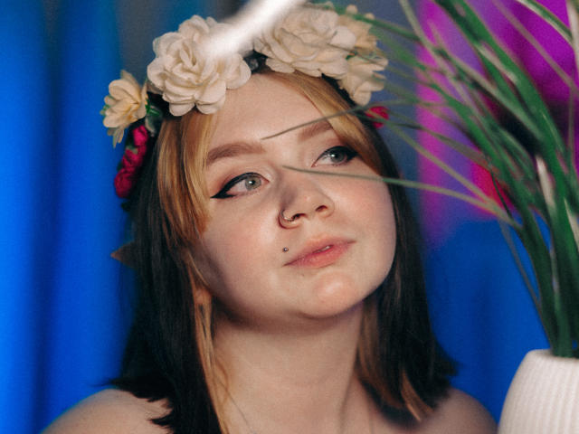 LunaLise with blonde and black hair, floral headband, nose piercing, and lip piercing, looking up, holding green plant leaves, blue background, soft light, slight blush on her cheeks.