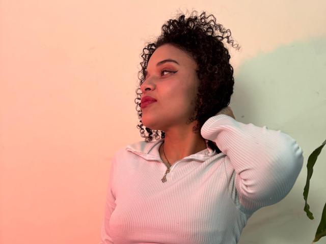 CattaleyaMolina with curly black hair, pink lipstick, and a white ribbed shirt, looking to the side with a hand in her hair, showing her tits slightly.
