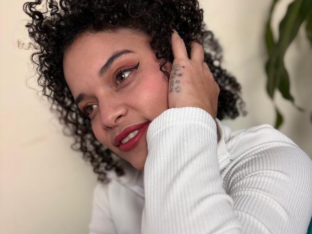 CattaleyaMolina with curly black hair, red lipstick, and a white ribbed shirt, touching her face with a tattoo on her hand, smiling softly. Her tits are not visible.
