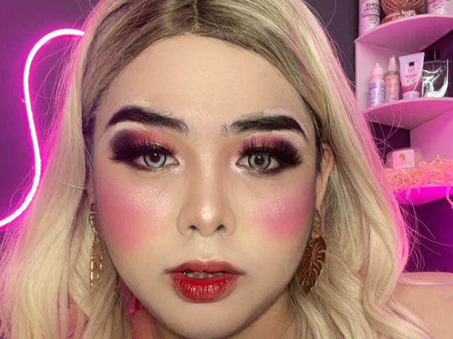 MisstresGela has blonde hair, pink blush, red lipstick, and dramatic eye makeup with thick lashes. She's wearing gold earrings and looking at the camera with a serious expression.