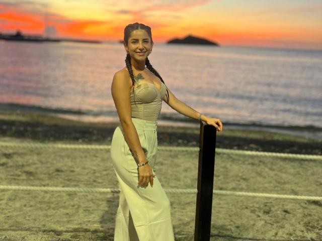 SofyaLane se tient sur une plage au coucher du soleil, vêtue d'une robe blanche sans manches avec une fente haute, ses mains sur un poteau noir, souriante, avec des cheveux tressés..