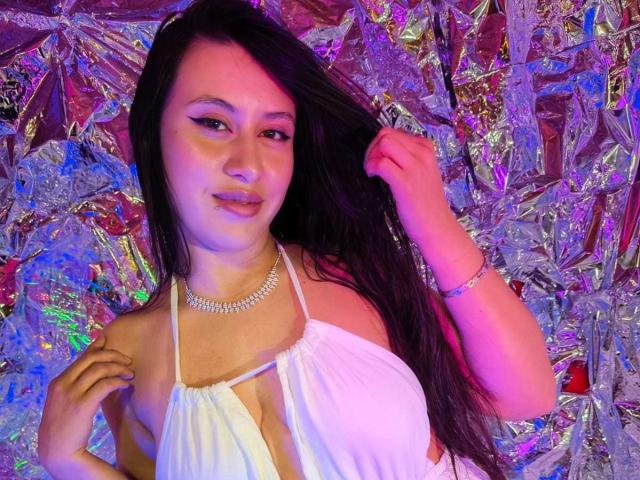 CeleneEvans with long black hair, white halter dress showing her tits, silver necklace, pink and blue light, holding hair, smiling, pink lipstick.