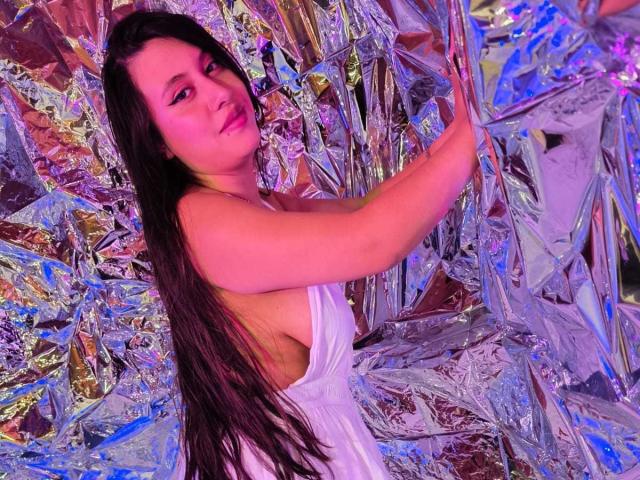 CeleneEvans with long dark hair, pink face light, wearing a low-cut white dress, touching shiny silver foil, looking at the camera, purple and blue lights in the background.