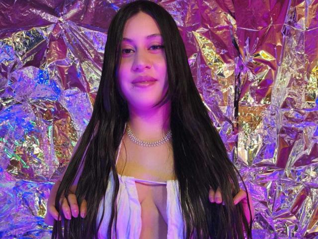 CeleneEvans with long black hair, purple and blue lighting, white open-cleavage top, holding hair, wearing a diamond necklace, smiling, shiny metallic background.