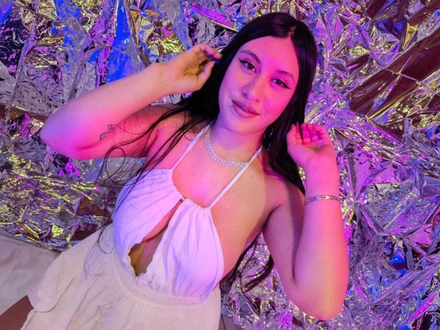 CeleneEvans with long black hair, white halter top, and visible cleavage, wearing a necklace and bracelet, touching her hair, smiling, pink and purple lighting, silver foil background.
