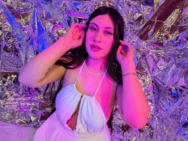 CeleneEvans with long black hair, wearing a white halter dress with cutout, showing her tits, pink and purple lighting, hands in her hair, silver necklace, bracelet, and a tattoo.