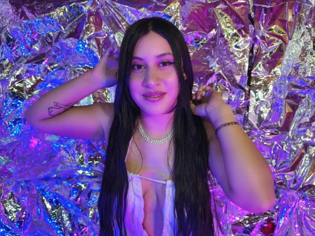 CeleneEvans with long black hair, pink eyeliner, and a pearl necklace, wearing a sheer top, hands in her hair, smiling, purple and blue lighting, tattoo on right arm.