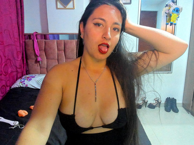 VenusSweetPalmer with long black hair, sticking out tongue, wearing a black halter top that shows her tits, hand in hair, necklace, and a pink room in the background.
