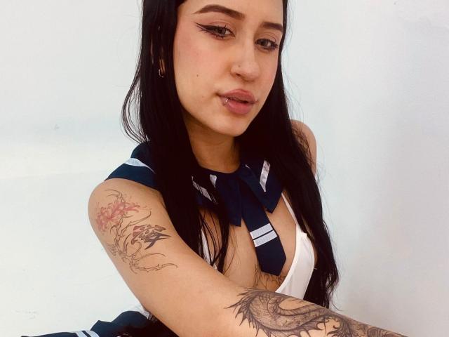 AnnaTorress with long black hair, eyeliner, and pink lips, wearing a navy and white schoolgirl outfit, showing tattooed arms, and a nose ring, looking at the camera.