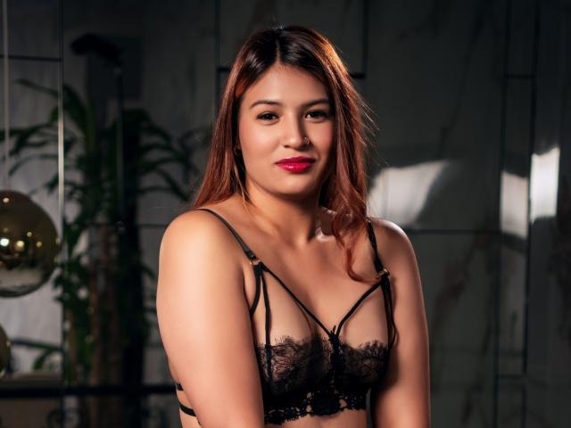 ZiaStorm, con piel marrón media, lápiz labial rojo y cabello castaño lacio, vestida con un sujetador de encaje negro, mostrando pequeños senos, de pie en una habitación con poca luz, sonriendo a la cámara..