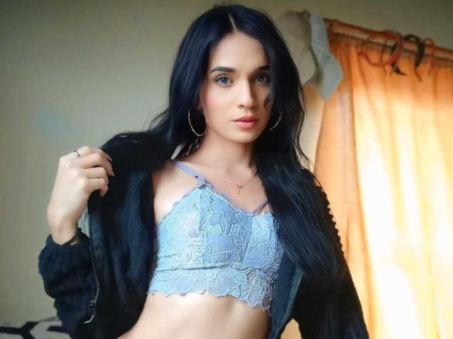 PamelaKander with long black hair, blue lace bra showing her tits, black jacket, gold hoop earrings, and a cross necklace, standing in front of orange curtains.