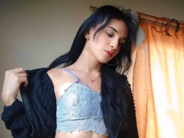 PamelaKander with long black hair, wearing a lace blue bralette, black jacket, and gold earrings, pulling jacket off, showing her tits, standing by orange curtains.