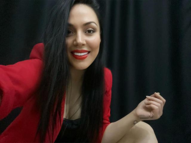 Layla84-sexy, com cabelos longos e pretos, lábios vermelhos e blazer vermelho, sentada e sorrindo, exibindo seus seios com um braço levantado e fundo preto..