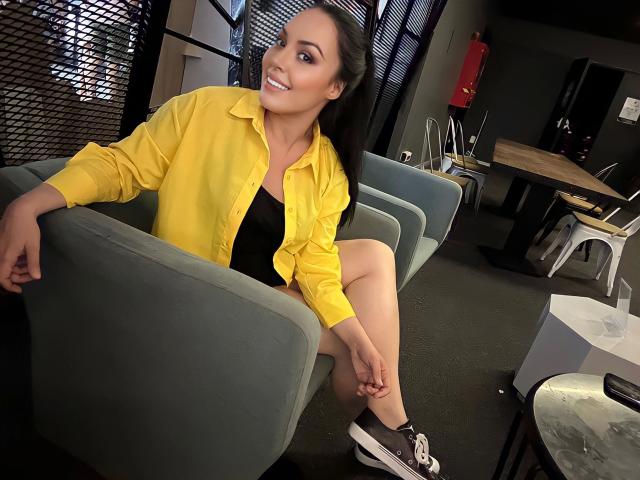 Layla84-hot with dark hair, yellow shirt, black top, and shorts, sitting on a blue chair, smiling, legs crossed, wearing black and white sneakers, in a dimly lit room.