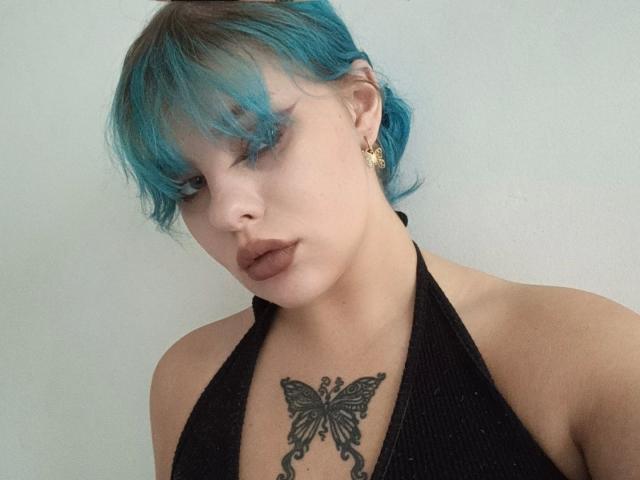 LunaGeil-hot a les cheveux bleus, un rouge à lèvres foncé et un tatouage de papillon sur la poitrine. Elle porte un haut noir à encolure dégagée, des boucles d'oreilles dorées et regarde vers le bas..